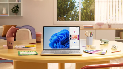 Portátil con Windows 11 en una mesa amarilla circular junto a materiales de escritorio en una habitación luminosa.