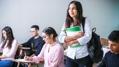 Una joven con mochila y cuaderno de pie en un aula mientras otros estudiantes toman apuntes sentados en sus pupitres.