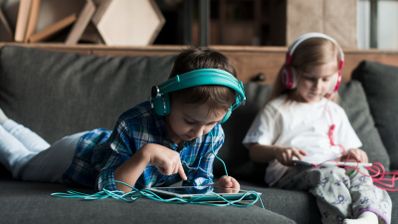 Niños en su propio hogar sosteniendo dispositivos móviles con auriculares puestos.