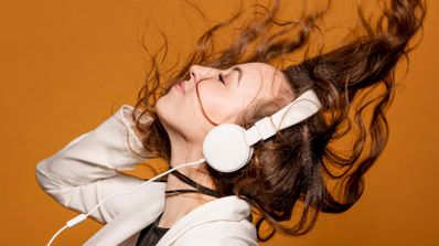 Mujer con auriculares blancos moviendo su melena castaña al ritmo de la música sobre un fondo ocre.