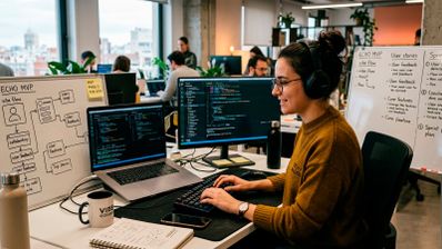 Una mujer en un entorno de oficina moderna, sonriendo mientras codifica en dos monitores con auriculares puestos. Tiene un ordenador portátil abierto y una taza de café en su escritorio, junto a una pizarra con diagramas.
