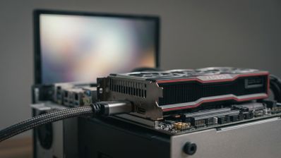 Primer plano de una tarjeta gráfica AMD Radeon conectada mediante un cable HDMI trenzado a un ordenador, con un monitor desenfocado al fondo.