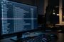 Monitor de ordenador en un entorno oscuro mostrando una lista de procesos del sistema con iconos de verificación y una alerta de seguridad resaltada en rojo.