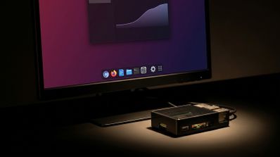 Monitor que muestra la interfaz de Raspberry Pi OS con un nuevo dock inferior de aplicaciones junto a una placa Raspberry Pi en su carcasa.