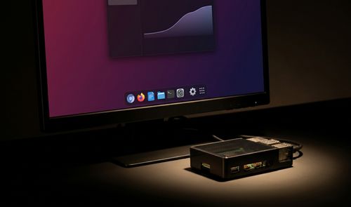 Monitor que muestra la interfaz de Raspberry Pi OS con un nuevo dock inferior de aplicaciones junto a una placa Raspberry Pi en su carcasa.