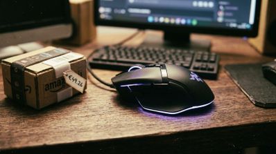 Ratón Razer Basilisk V3 con iluminación RGB sobre un escritorio de madera junto a un paquete de Amazon con una etiqueta de precio.