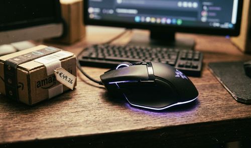 Ratón Razer Basilisk V3 con iluminación RGB sobre un escritorio de madera junto a un paquete de Amazon con una etiqueta de precio.