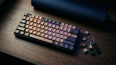 Teclado mecánico gaming Logitech con retroiluminación RGB en tonos cálidos y violetas sobre una mesa de madera, junto a teclas de repuesto y una herramienta extractora.