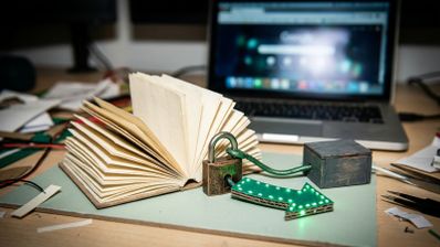 Libro abierto junto a un candado y una flecha con luces LED verdes en una mesa de trabajo con un ordenador portátil al fondo.