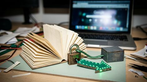 Libro abierto junto a un candado y una flecha con luces LED verdes en una mesa de trabajo con un ordenador portátil al fondo.