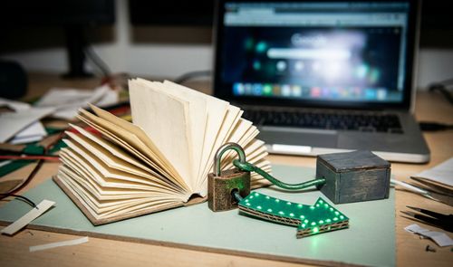 Libro abierto junto a un candado y una flecha con luces LED verdes en una mesa de trabajo con un ordenador portátil al fondo.