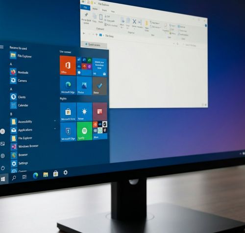 Monitor de ordenador mostrando el escritorio con el menú Inicio clásico de baldosas y una ventana del Explorador de archivos abierta sobre un fondo degradado azul y violeta.