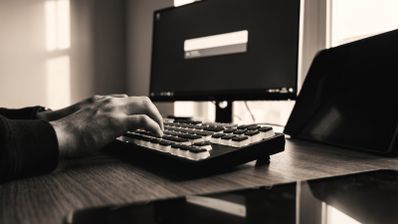 Manos de una persona utilizando un teclado mecánico frente a un monitor que muestra la interfaz de búsqueda de Flow Launcher en Windows.