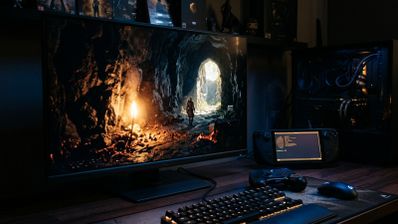Monitor Dell mostrando un videojuego en una cueva iluminada junto a una Steam Deck con una terminal de comandos abierta sobre un escritorio.