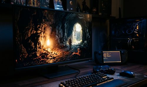 Monitor Dell mostrando un videojuego en una cueva iluminada junto a una Steam Deck con una terminal de comandos abierta sobre un escritorio.