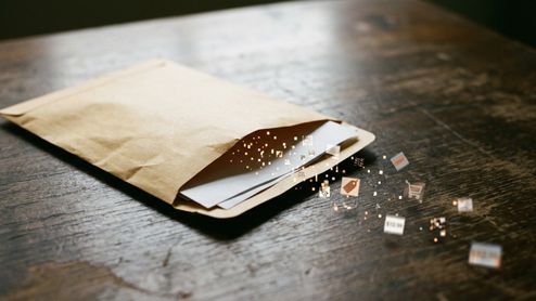 Sobre de papel abierto en una mesa de madera del cual salen partículas digitales e iconos de compras y precios.
