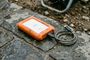Disco duro externo LaCie Rugged Mini con funda protectora naranja sobre suelo de piedra mojado.