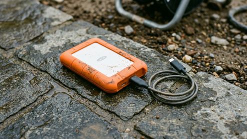 Disco duro externo LaCie Rugged Mini con funda protectora naranja sobre suelo de piedra mojado.