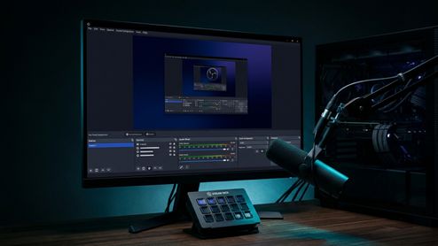 Interfaz de OBS Studio abierta en un monitor junto a un micrófono profesional, una consola Stream Deck y un PC de sobremesa.