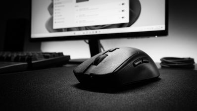 Primer plano de un ratón de ordenador negro sobre una alfombrilla frente a un monitor con Windows 11.