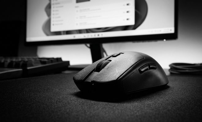 Primer plano de un ratón de ordenador negro sobre una alfombrilla frente a un monitor con Windows 11.