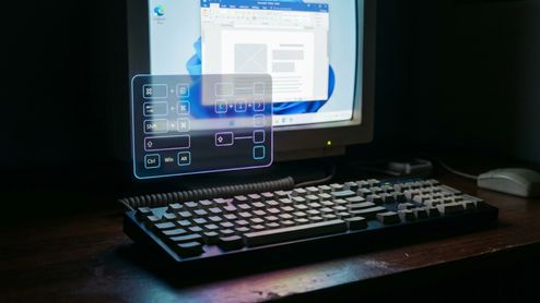 Teclado mecánico clásico en primer plano frente a un monitor con una interfaz de realidad aumentada que muestra diagramas de atajos de teclado.
