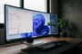 Monitor ultra ancho con la interfaz de Windows 11 mostrando el explorador de archivos y el menú de inicio sobre un escritorio de madera.